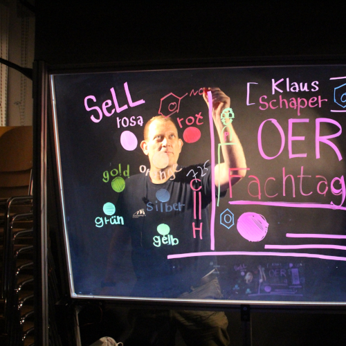 Foto von Klaus Schaper, der auf ein Lightboard schreibt. Das Lightboard wird in pink beschrieben, es sind Wörter zu lesen: Klaus Schaper, OER Fachtag, Sell
